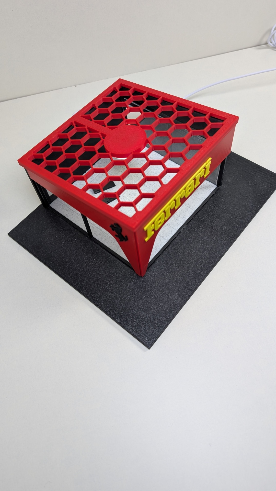 Miniatura estrutura cúbica com base preta e topo vermelho hexagonal com texto FERRARI amarelo.