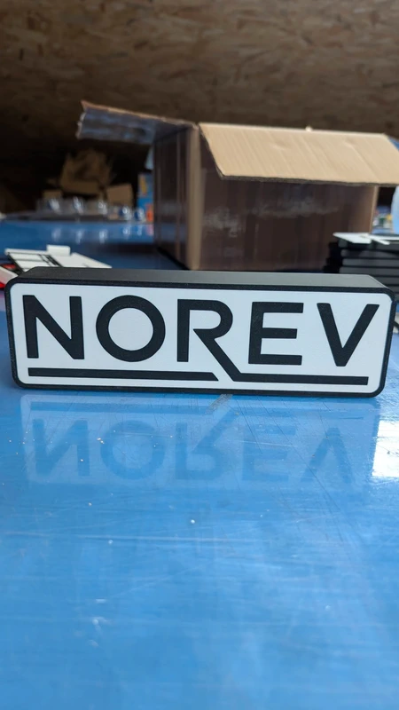 Placa com texto Norev em fundo branco e letras pretas sobre superfície azul reflexiva.