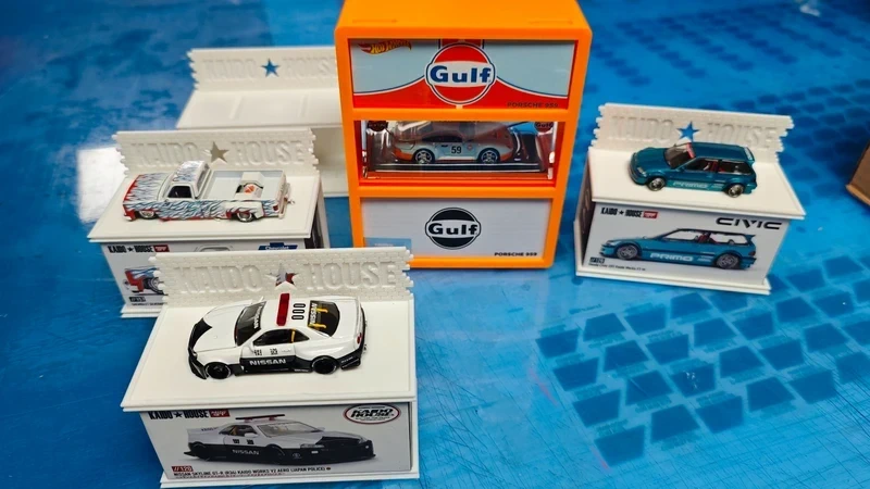 Quatro carros em miniatura em suportes brancos e caixa laranja Gulf sobre mesa azul
