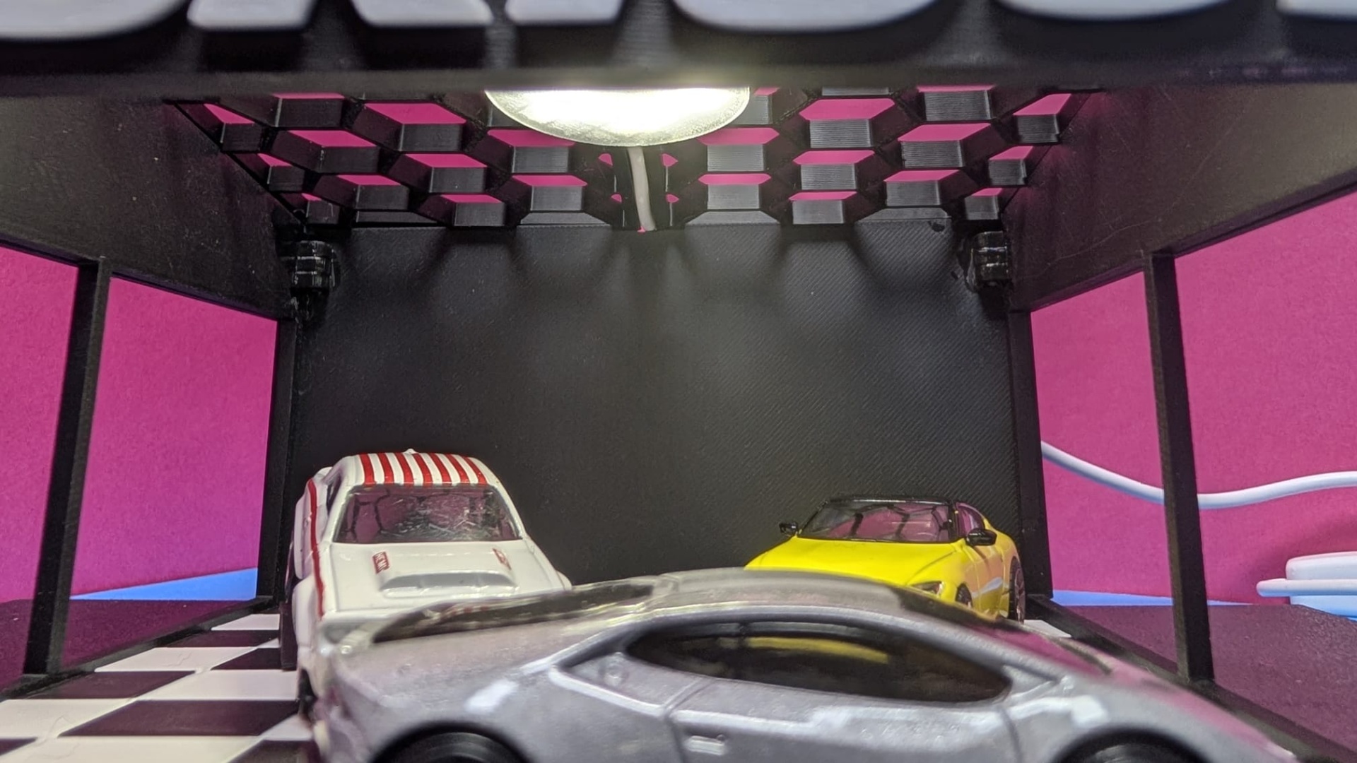 Três carros de brinquedo numa garagem preta com chão xadrez e painéis cor de rosa nas laterais.