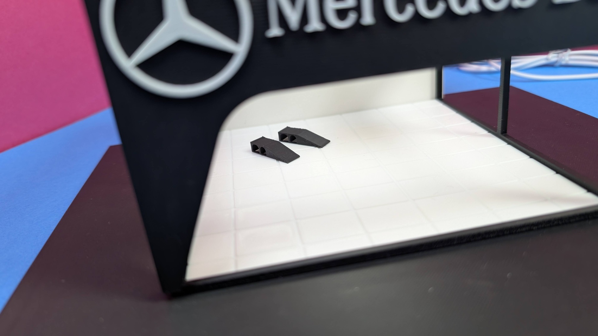 Base decorativa com logótipo da Mercedes-Benz e duas pequenas peças triangulares pretas em superfície branca.