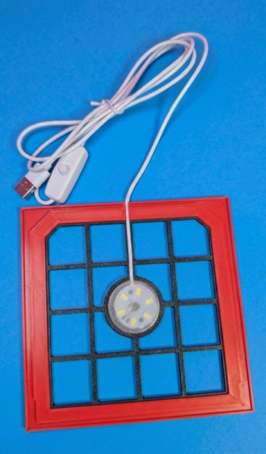 Tapete quadrado vermelho com grelha preta, círculo central com LEDs e cabo USB branco com interruptor.
