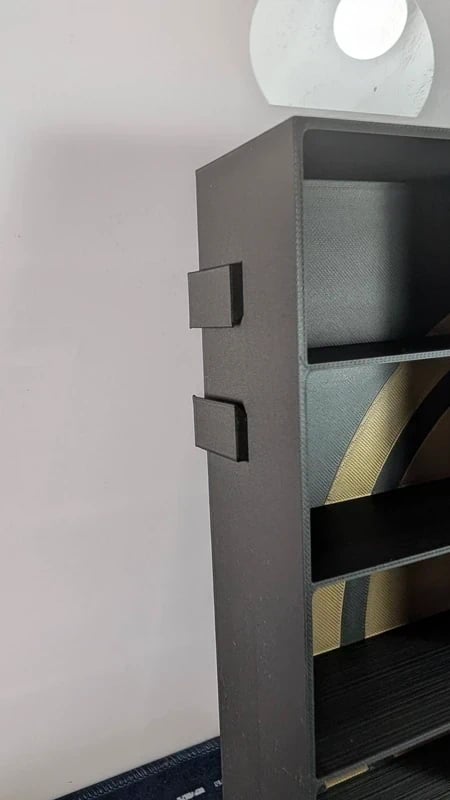 Prateleira preta aberta com objeto circular branco no topo e padrão geométrico dourado no fundo