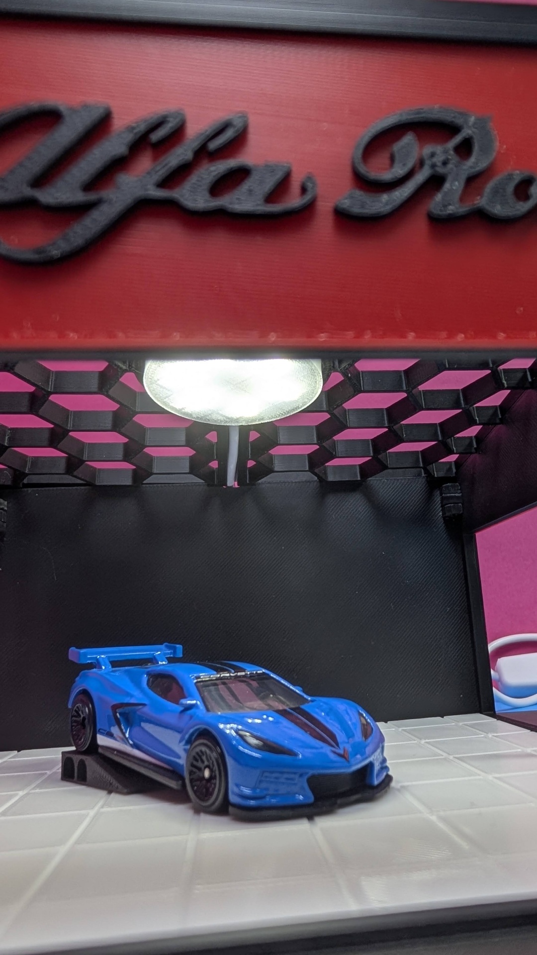 Miniatura de carro desportivo azul em suporte preto sobre base branca, fundo preto e vermelho com letras pretas.