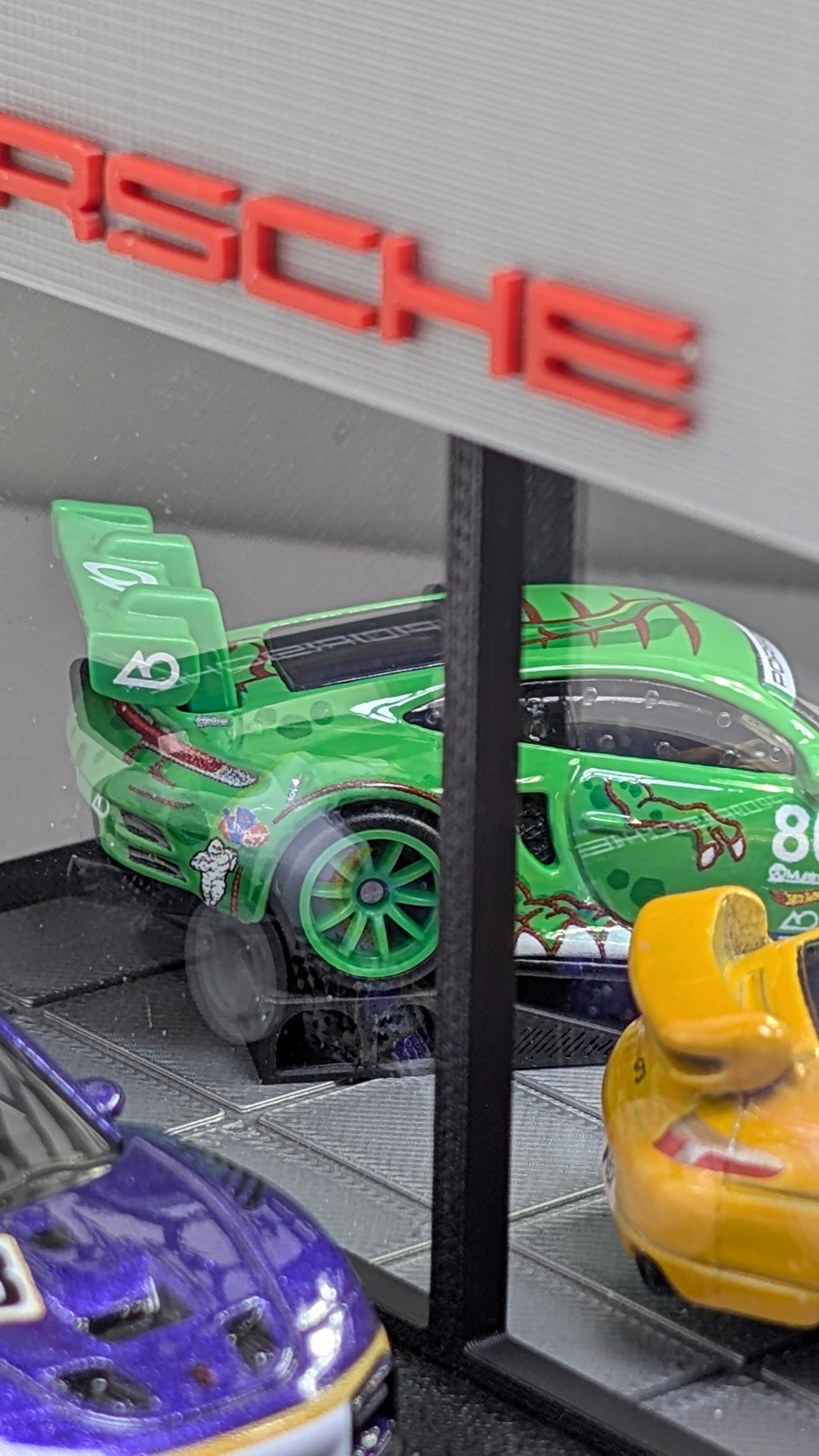 Carros de brinquedo Porsche em miniatura verdes, amarelos e roxos em vitrina com logo vermelho