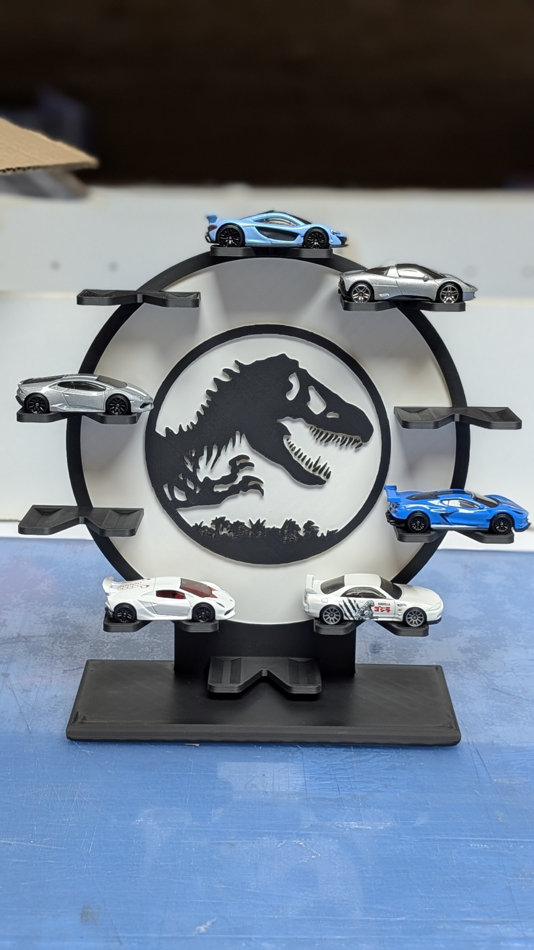 Expositor circular preto com mini carrinhos de metal coloridos e desenho de dinossauro ao centro