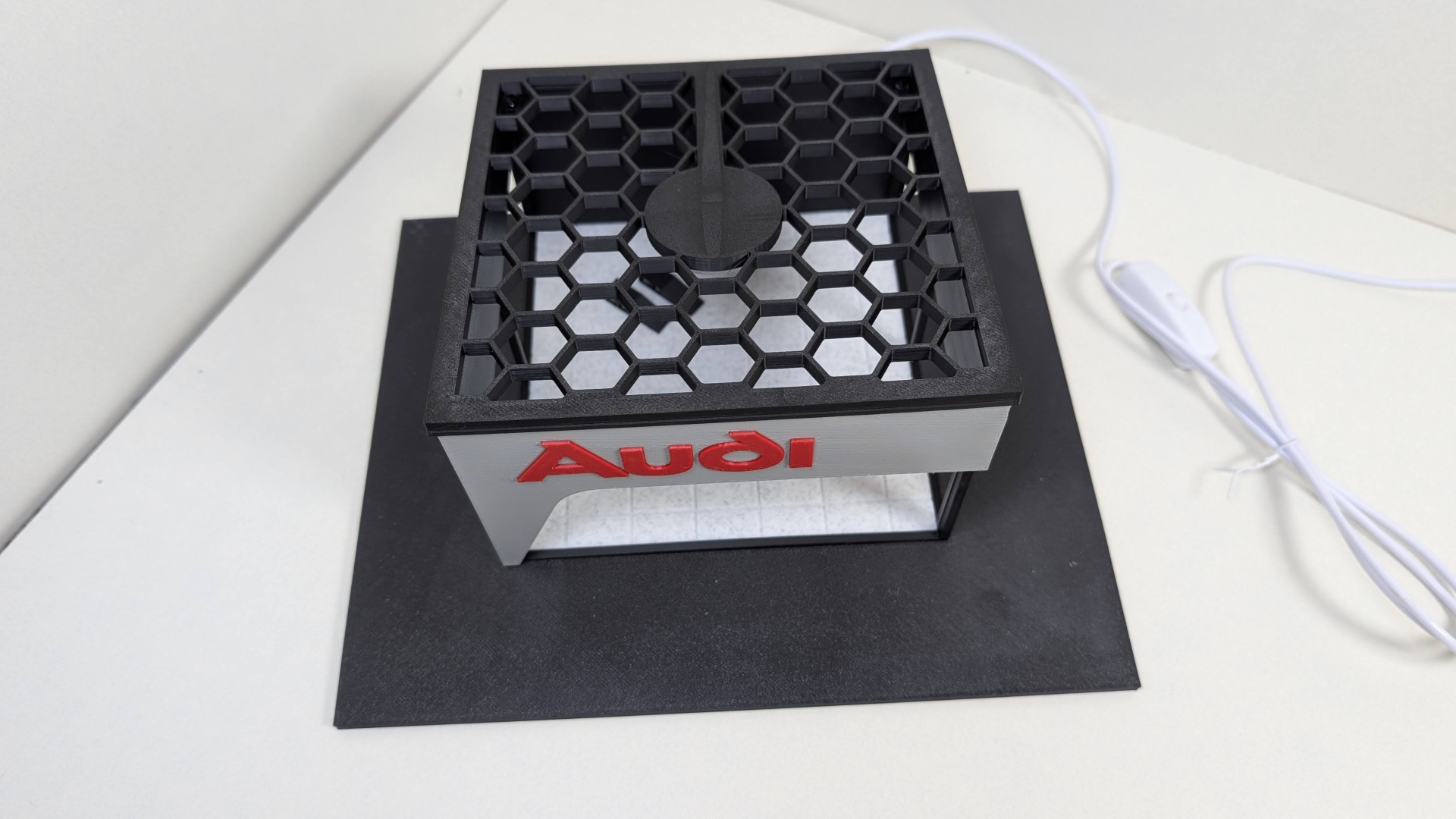 Objeto com padrão hexagonal e texto Audi vermelho numa base preta com cabo branco