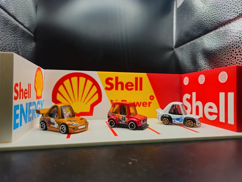 Três carrinhos de brinquedo em miniatura numa superfície com marcações vermelhas e fundo publicitário da Shell