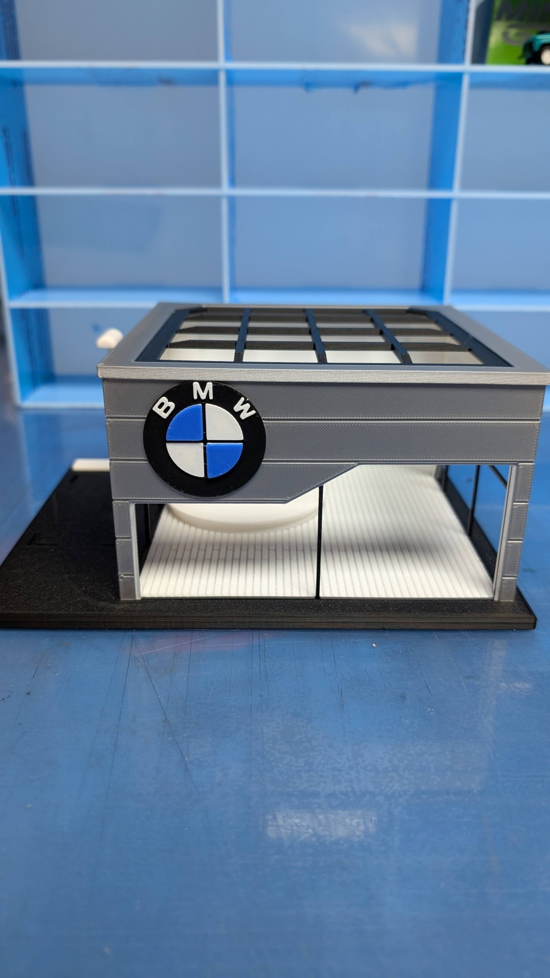 Miniatura de prédio cinzento com logótipo BMW e estante azul no fundo