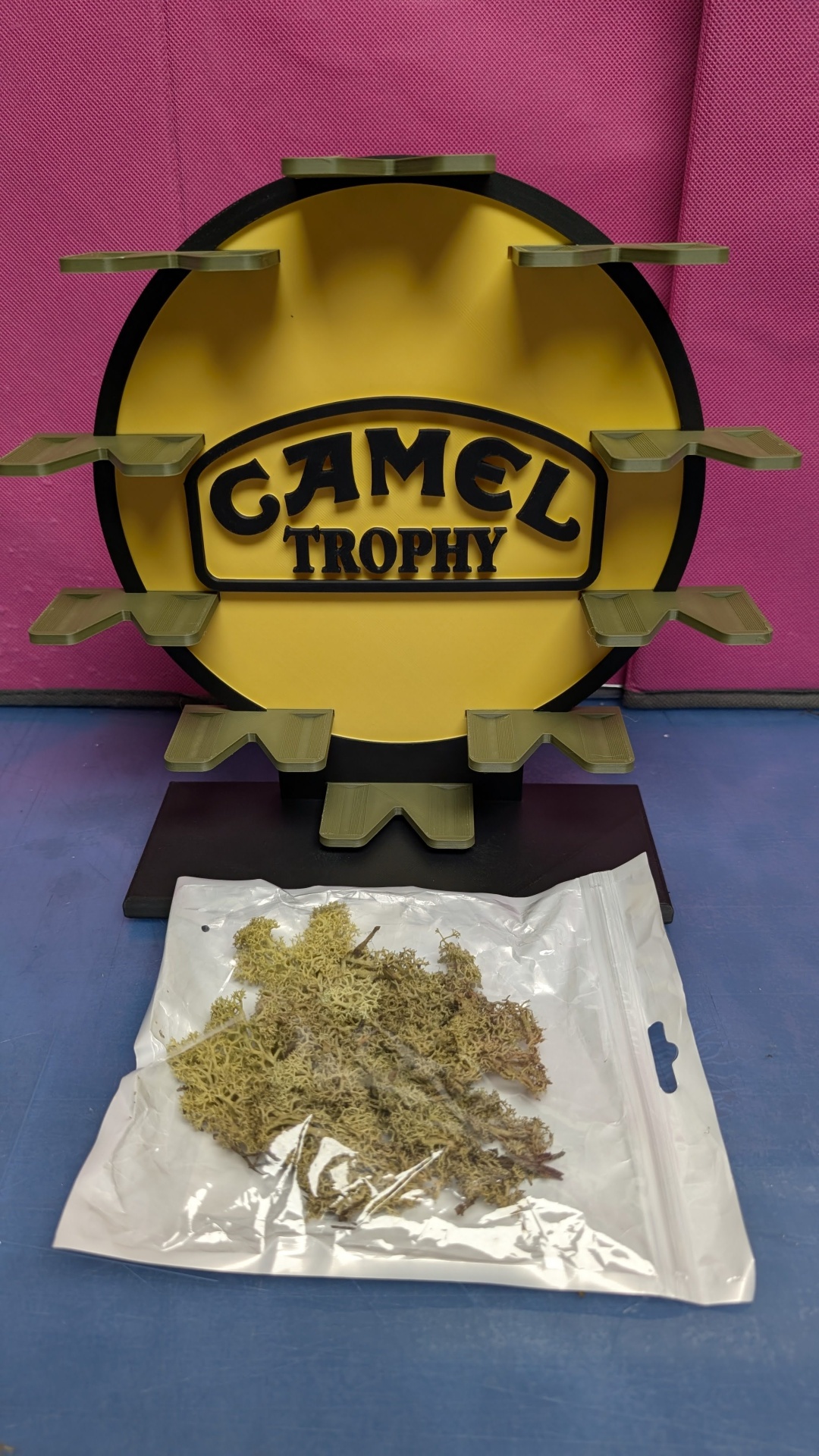 Expositor CAMEL TROPHY amarelo e preto com prateleiras verdes e saco plástico com musgo seco