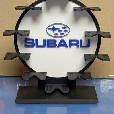 Suporte circular com logótipo da Subaru azul sobre fundo branco e base preta.