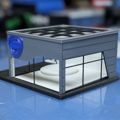 Maqueta de edifício com estrutura cinzenta e janelas de vidro, logotipo azul LANCIA