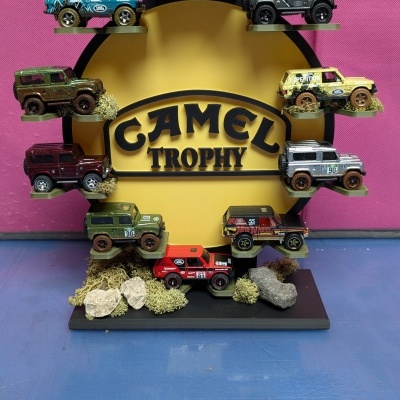 Miniaturas de carros Land Rover exibidas em suporte CAMEL TROPHY com decoração natural.