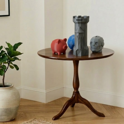 Mesa redonda de madeira com porquinho mealheiro vermelho, torre cinzenta, esfera azul e cabeça cinzenta decorativas num canto de parede clara ao lado de planta em vaso