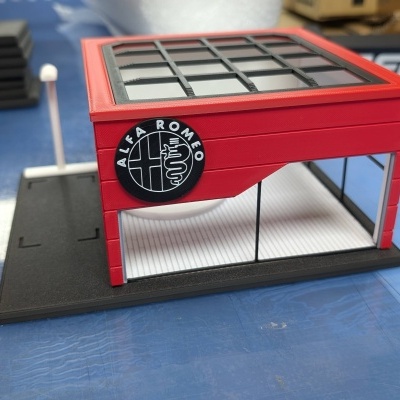 Miniatura de prédio vermelho com janelas de vidro e logo ALFA ROMEO em fundo azul