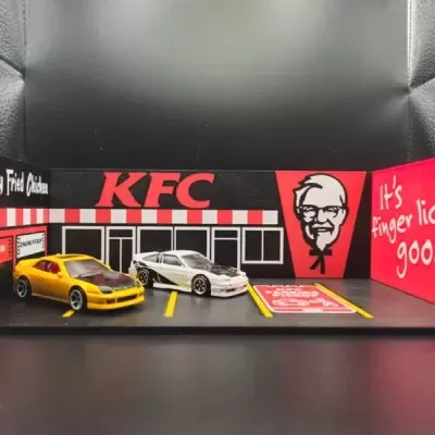 Miniaturas de carros num parque de estacionamento miniatura com fachada da KFC e texto promocional