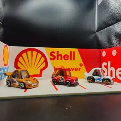 Três carrinhos de brinquedo em miniatura numa superfície com marcações vermelhas e fundo publicitário da Shell