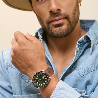 Relógio masculino de mostrador preto com pulseira de pele castanha e detalhes amarelos no pulso de homem de camisa de ganga azul.