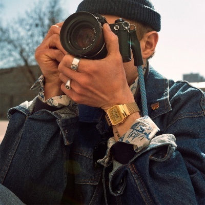 Homem com casaco e camisa azul a usar máquina fotográfica preta com lente 50mm e relógio digital dourado