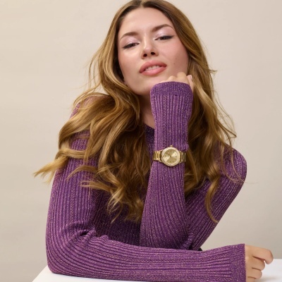 Relógio dourado com pulseira metálica no pulso de mulher com camisola roxa brilhante