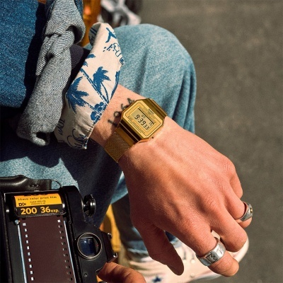 Relógio digital Casio dourado no pulso de uma pessoa segurando uma câmara fotográfica analógica.