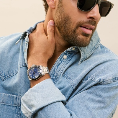 Relógio de pulso prateado com mostrador azul em homem com camisa de ganga e óculos escuros