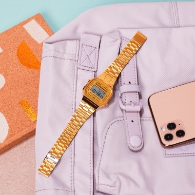 Relógio digital dourado com bracelete metálica sobre mochila lilás, telemóvel dourado e embalagem laranja com texto