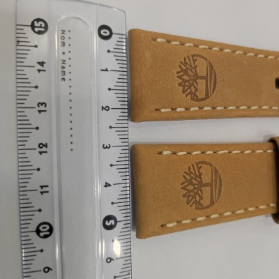 Duas tiras de pulseira em couro castanho claro com costura branca e logótipo Timberland ao lado de régua transparente