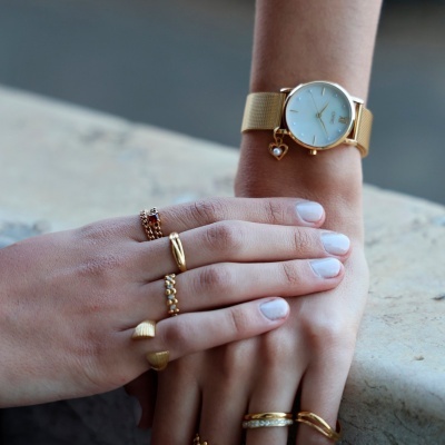 Relógio dourado com mostrador branco e bracelete metálica, vários anéis dourados e prateados na mão