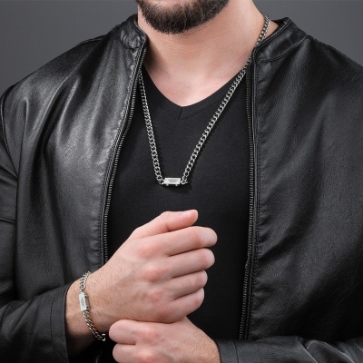 Homem com colar e pulseira metálicos em roupa preta e fundo cinzento