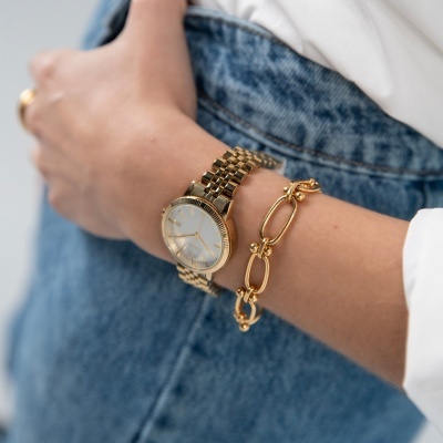 Relógio de pulso dourado com pulseira e pulseira dourada ao lado, no pulso de pessoa com camisa branca e jeans azuis