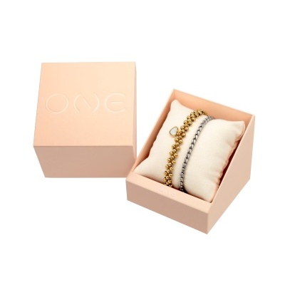 Conjunto de pulseiras dourada e prateada numa almofada branca dentro de caixa rosa com texto OME