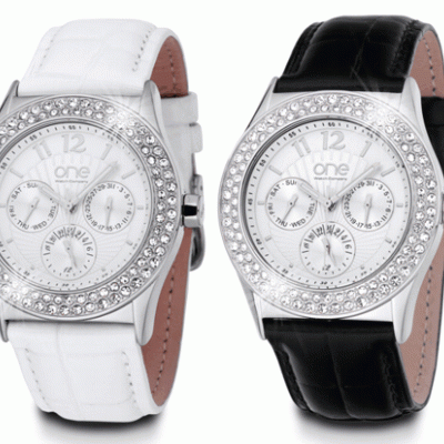 Dois relógios de pulso com braceletes em couro, um branco e outro preto, mostradores redondos prateados com pedras brilhantes.