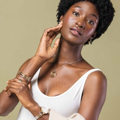 Mulher com colar, pulseiras douradas, blusa branca de alças finas e casaco bege