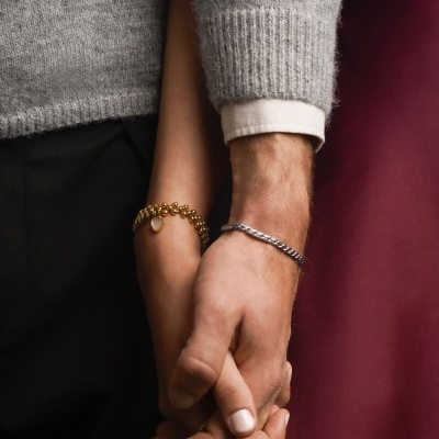 Dois braços entrelaçados com pulseiras dourada e prateada, roupas cinza, preto e bordeaux.