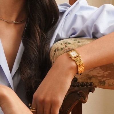 Relógio dourado de pulso com mostrador retangular usado por mulher vestindo camisa azul clara sentada numa cadeira estofada