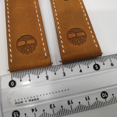 Correias de couro castanho com costura branca e logótipo gravado de árvore sobre régua transparente