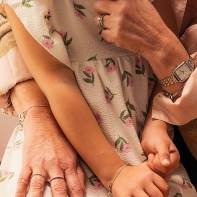 Vestido infantil branco com padrão floral rosa e verde, criança com joias prateadas e adulto com relógio e anéis