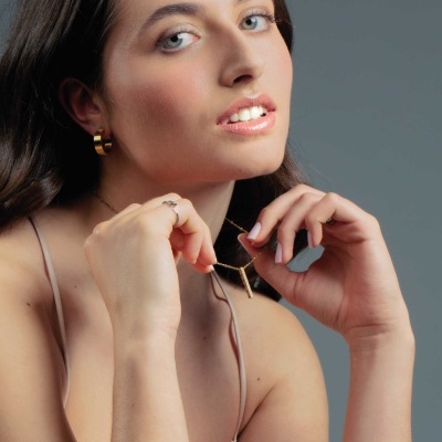 Mulher usando brincos dourados, colar e anéis, com top bege e fundo cinza