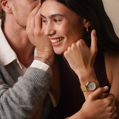 Relógio dourado com bracelete metálica numa mulher, pulseira e brincos dourados, ambiente interior creme