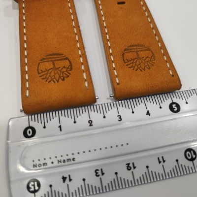 Bandas de relógio castanhas de couro com costura branca e logótipo de árvore sobre régua transparente.