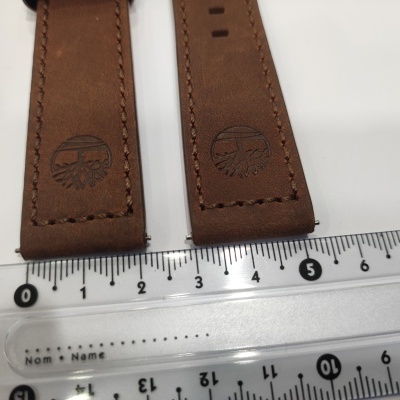 Duas pulseiras de relógio em couro castanho com logotipo gravado, réguas transparentes ao lado.