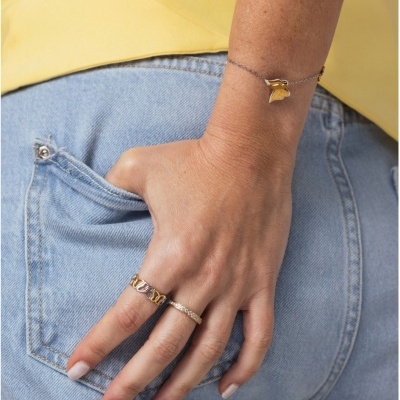 Mão feminina com anéis dourados e pulseira com pendente, calças jeans azuis e camiseta amarela.