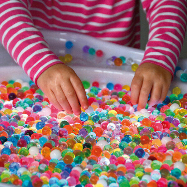 Mãos de criança com camisola às riscas a tocar esferas coloridas translúcidas numa bandeja branca.