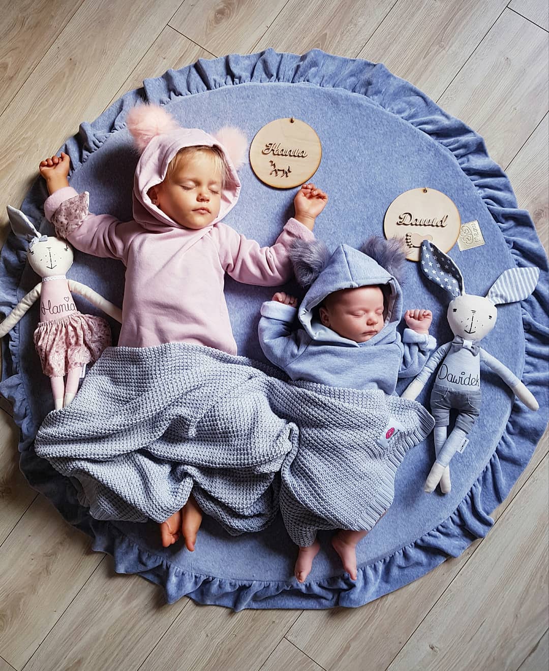Playmat MeowBaby® Dois bebés com roupas azuis e rosas a dormir numa manta azul redonda com folhos, rodeados por bonecas de pano e placas com nomes.