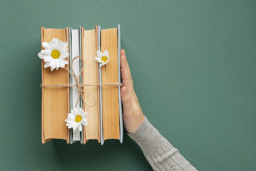 Mão segurando três livros amarrados com cordão e decorados com flores brancas em fundo verde