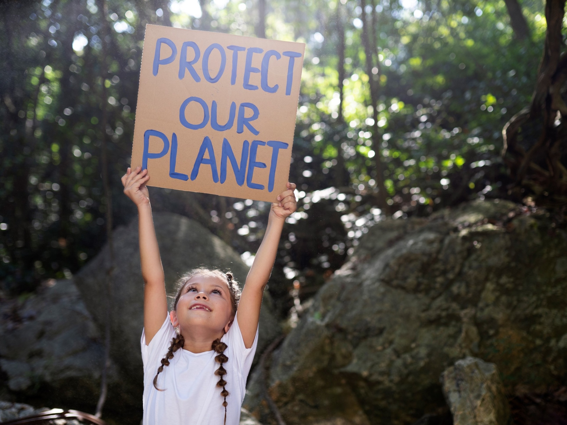 Criança numa floresta a segurar cartaz com texto 'PROTECT OUR PLANET'.