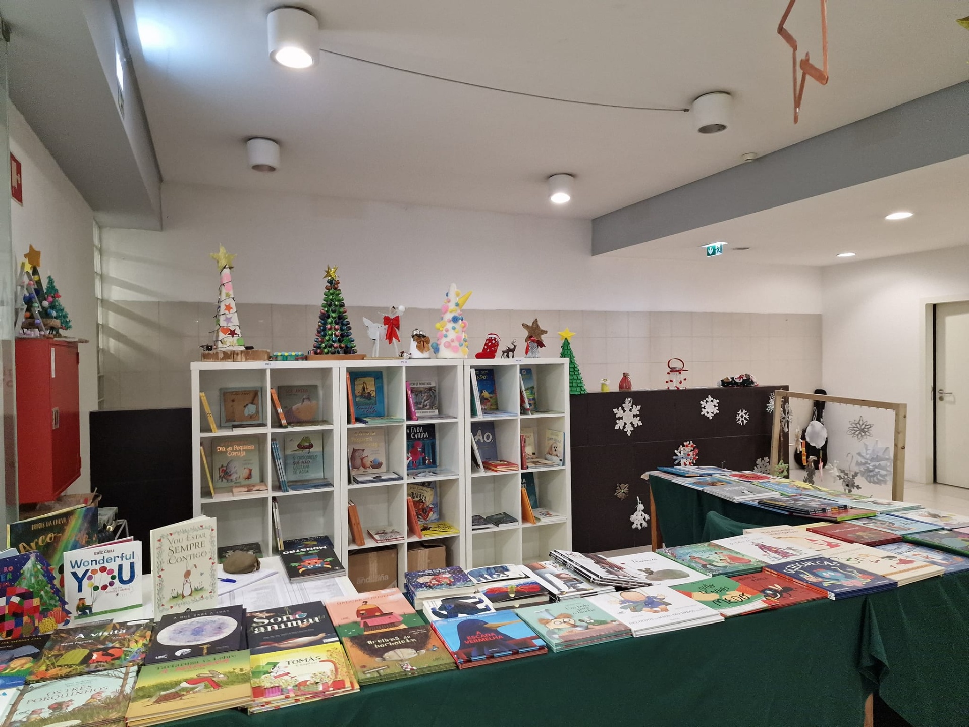 Mesa de feira com livros infantis e decorações natalícias em estantes brancas num espaço interior