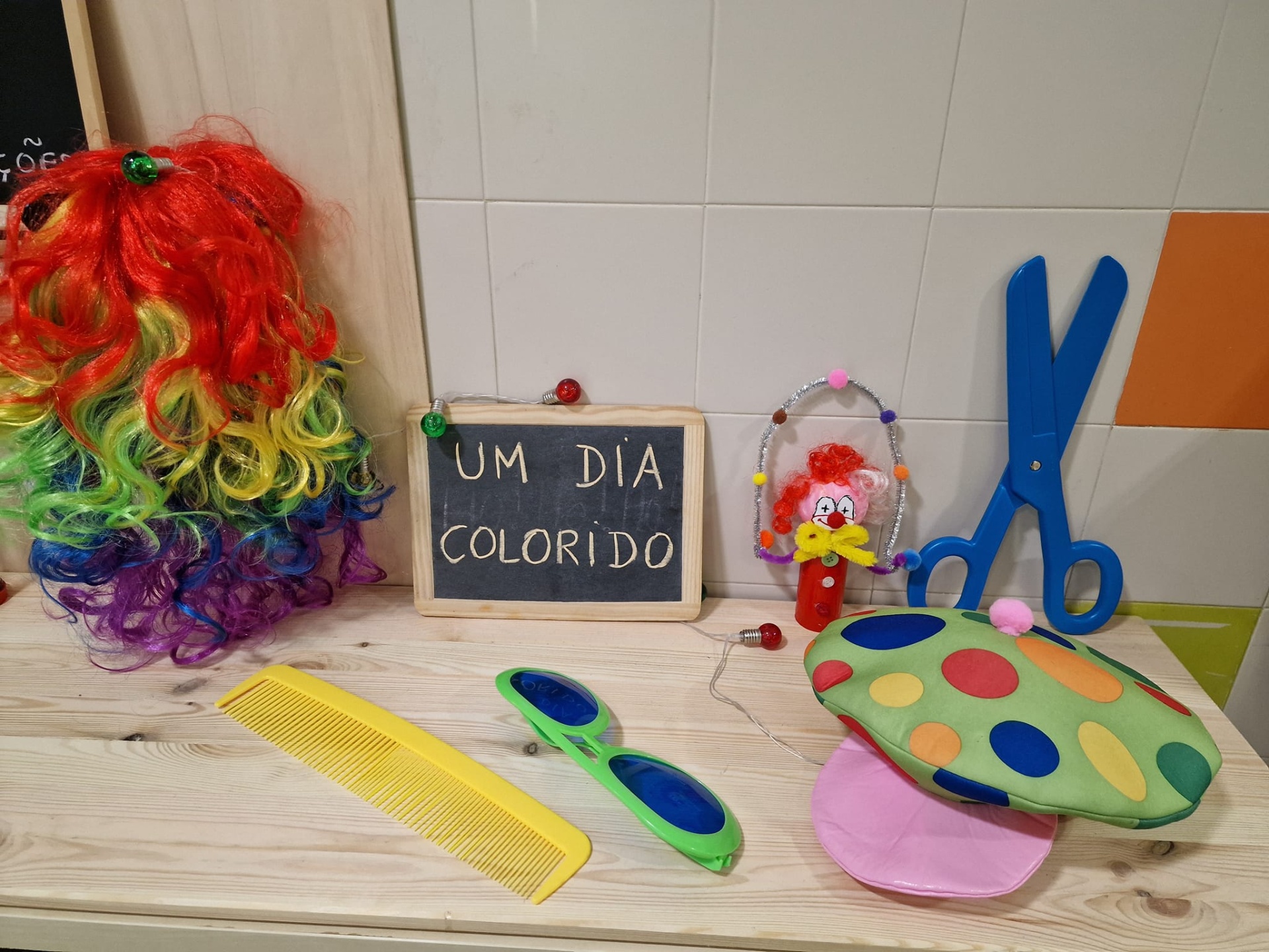 Acessórios coloridos para disfarce com peruca, chapéu e óculos sobre bancada com fundo de azulejos