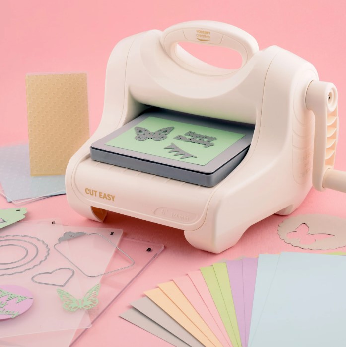 Cut Easy Cutting and Embossing Machine A5 Starter Kit Máquina de corte manual branca CUT EASY com papéis coloridos e moldes transparentes sobre superfície rosa